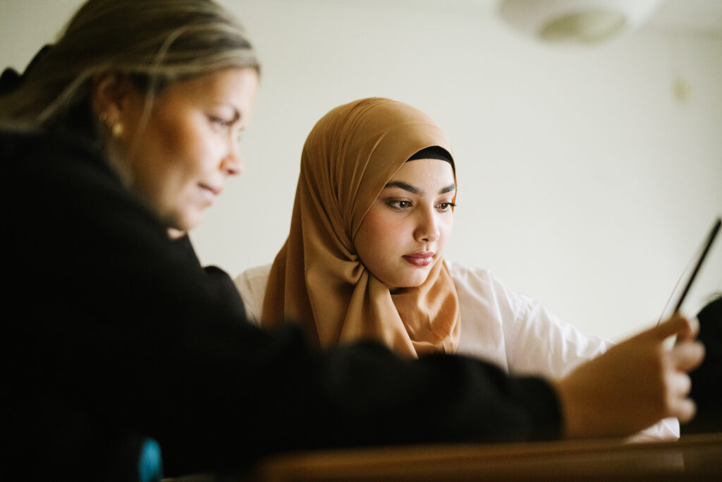 Två personer sitter framför en dator och båda tittar på skärmen. Personen i bakgrunden är i fokus i bild, medan personen i förgrunden sträcker sig mot datorskärmen.