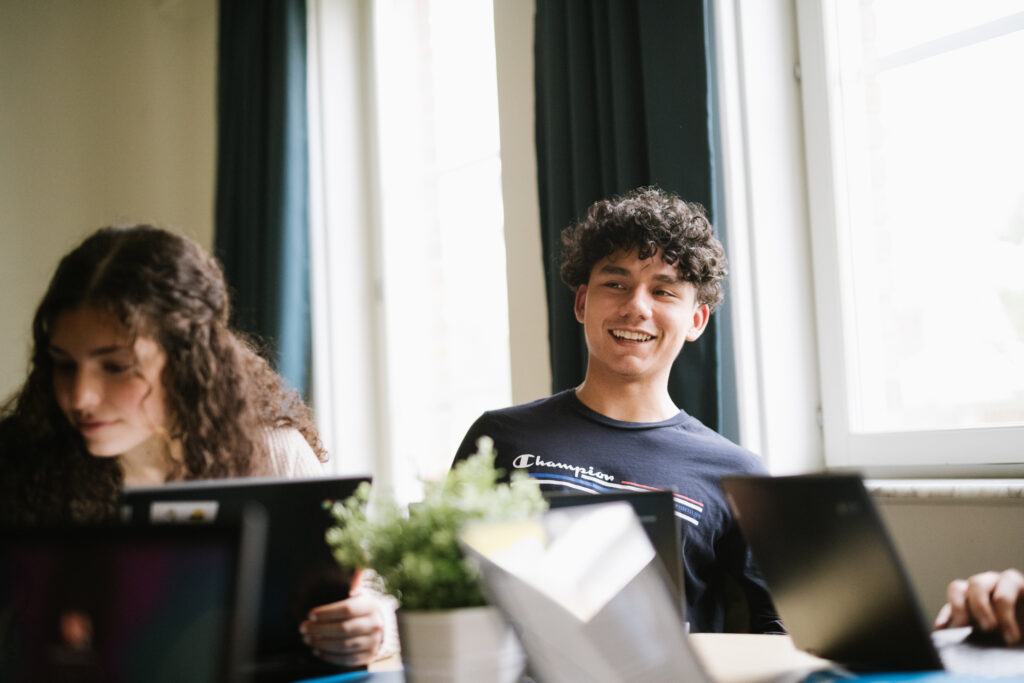 Två elever sitter bredvid varandra med varsin datorskärm i ett klassrum. En av eleverna är i fokus på bilden och sitter med ryggen mot ett fönster och skrattar