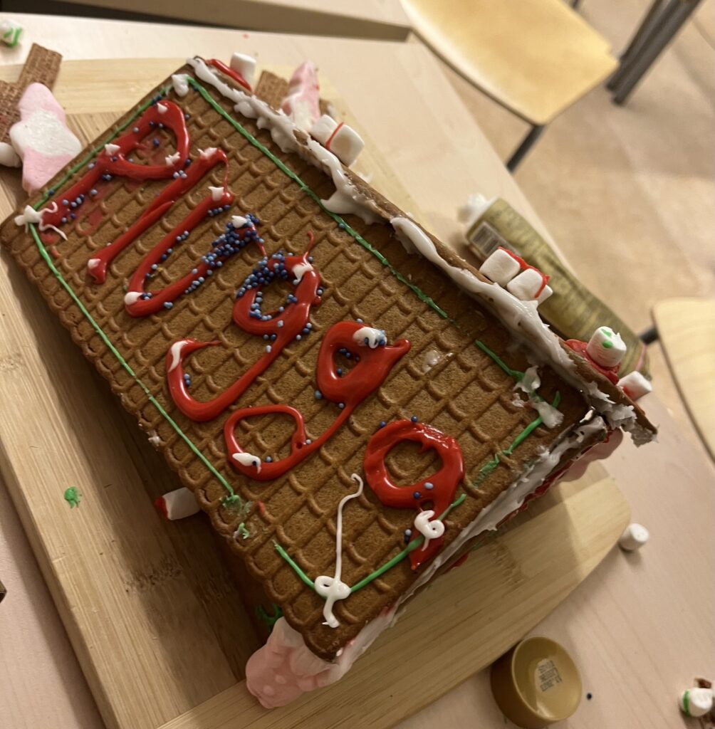 Johannesskolans TT1-grupp (Malmö). Bilden föreställer ett pepparkakshus med "Plugga" skrivet i röd glasyr på takets ena del, delen har även grön glasyr runt dess kanter. Vit glasyr ligger på toppen av taket. Nedanför taket står där en skum-tomte. Vid huset ligger även en tub glasyr.