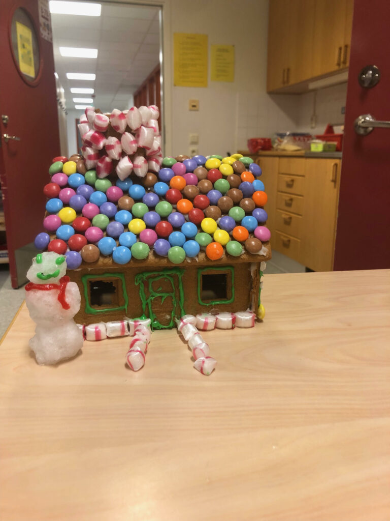 6. Nybyggeskolan TT1. Bilden föreställer ett pepparkakshus på ett bord. Pepparkakshuset har nonstop-choklad i olika färger som täcker hela taket. Skorstenen är beklädd med polkagris-karameller. Kanterna till fönstren och dörren på pepparkakshus har grön glasyr. Framför huset finns en gång beklädd med polkagris-karameller samt en snögubbe med röd halsduk av glasyr, samt ögon och mun med grön glasyr.