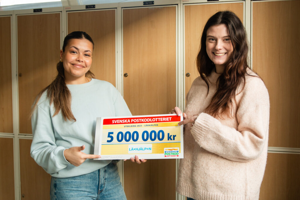 Läxhjälparen Sara och coach Greta framför skåp i en skolkorridor. De ler mot kameran och håller upp fem miljoners-checken.