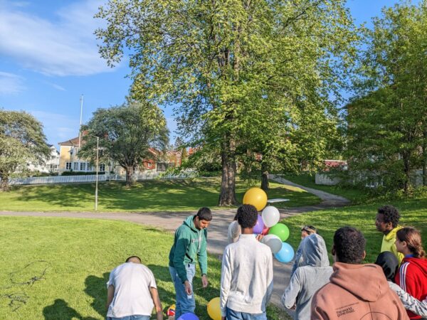 Sommaravslutning med grönskande träd och gräsmatta. I bild syns elever stå i grupp, med ryggarna mot kameran, och med ballonger i bild.