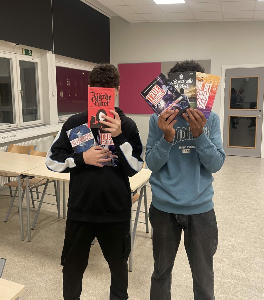 Två elever står i ett klassrum och håller upp böcker framför sina ansikten.