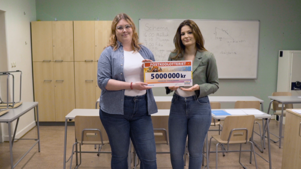 Två läxhjälpare står i ett klassrum och håller fram en check framför sig. På checken syns summan på fem miljoner kronor. Nedan syns Läxhjälpens logotyp och Svenska Postkodlotteriets logotyp. Bakom personerna finns en whiteboard-tavla där någon har skrivit ner ett schema för dagen.